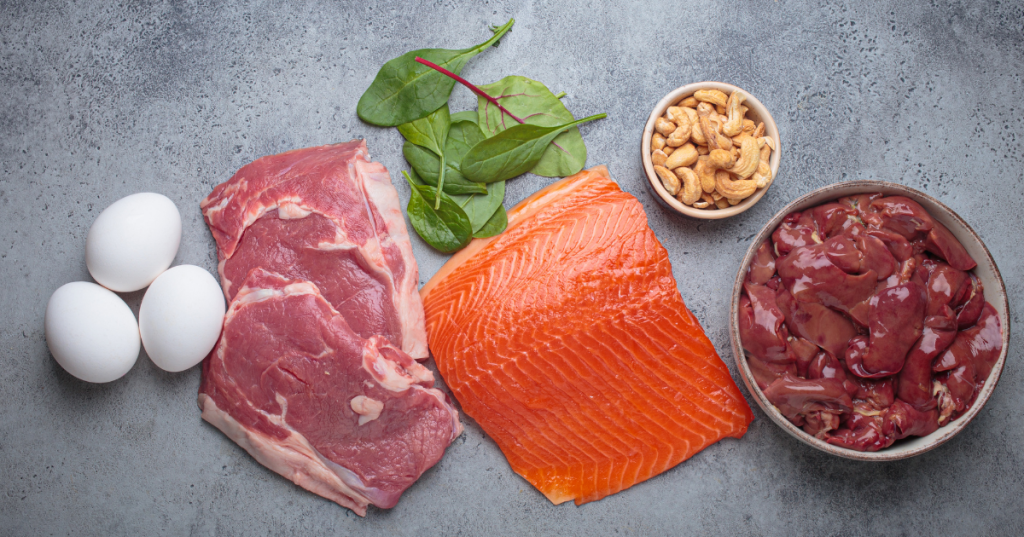 Assorted iron-rich foods on a gray background, including raw red meat, salmon, liver, eggs, spinach, and cashews, showing a mix of animal- and plant-based sources of iron.